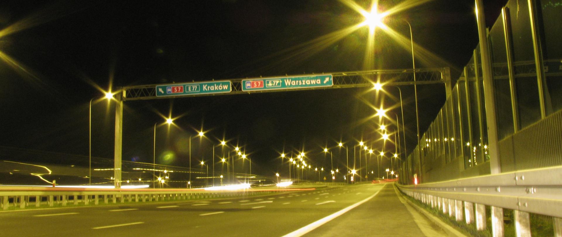 węzeł Kielce Północ - zdjęcie wykonane nocą, jezdnia dwujezdniowej drogo ekspresowej, z prawej strony jezdni ekrany, z lewej bariery, po obu stronach jezdni świecą się latarnie, blask lamp w kształcie gwiazd, nad jezdnią tablice kierunkowe z napisami "Kraków" i "Warszawa".
