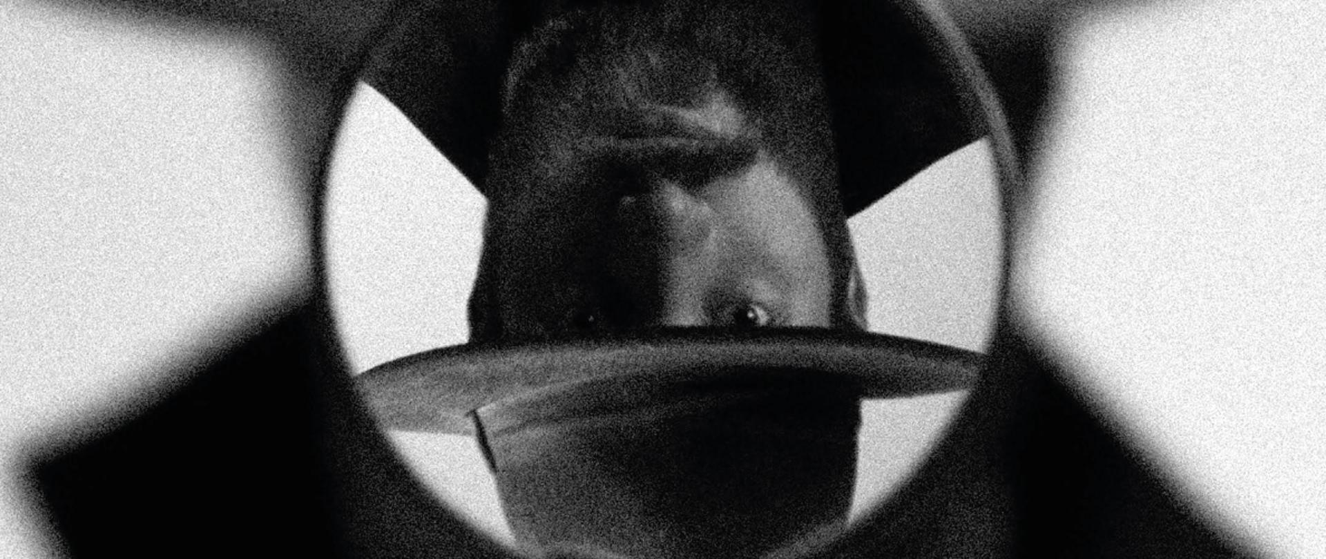 Zdjęcie w odcieniach szarości. Mężczyzna w kapeluszu w lupie odwrócony o 180 stopni.