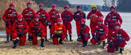 Strażacy Państwowej Straży Pożarnej zabezpieczeni w ubrania wypornościowe, kaski na głowie oraz liny ćwiczą na Jeziorze Dużym w Turawie działania związane z ratownictwem lodowym. Strażacy doskonalą techniki poruszania się po lodzie, samo ratownictwo z załamanego lodu oraz ewakuację osób poszkodowanych przy zastosowaniu sprzęty ratowniczego. Wspólne zdjęcie grupowe strażaków. Funkcjonariusze PSP stoją na plaży.