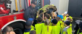 Dzieci stoją obok samochodu strażackiego i trzymają w górze kurtkę strażacką, a strażak opowiada o sprzęcie.