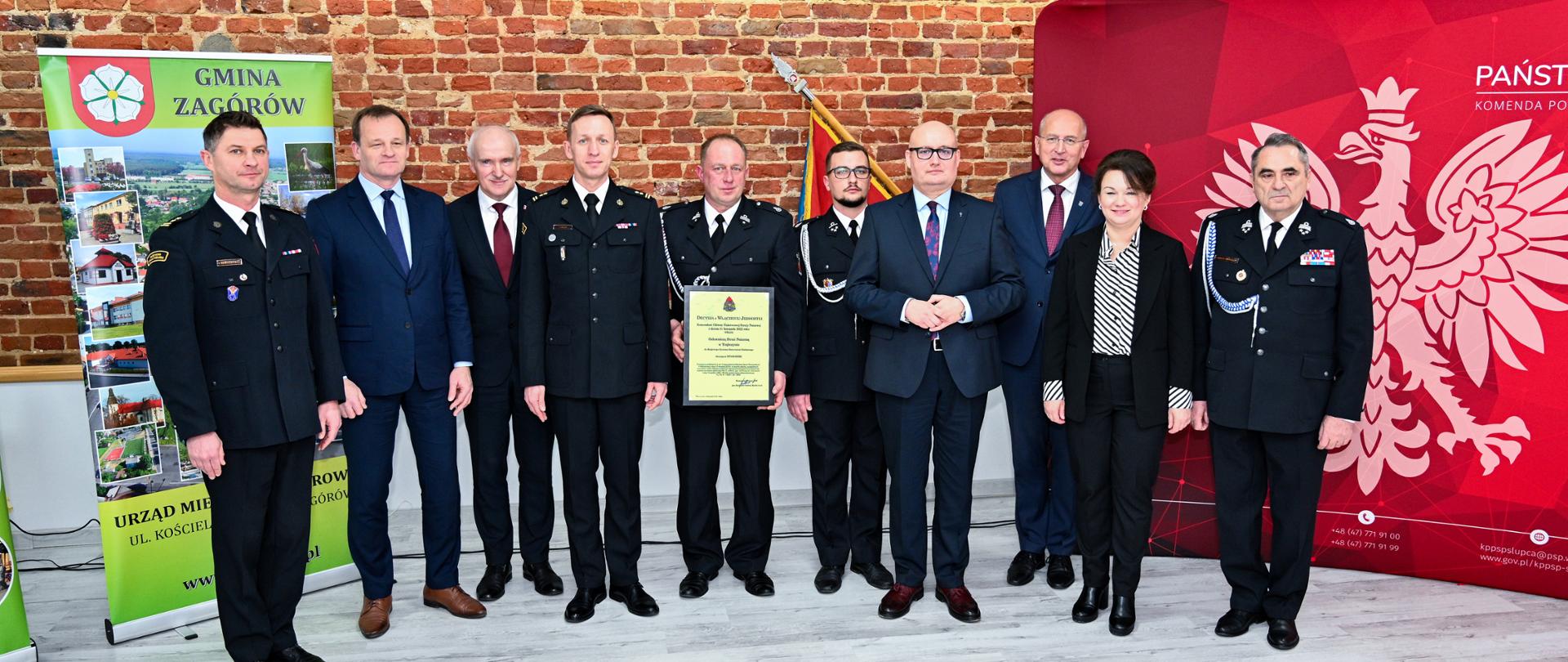 na zdjęciu funkcjonariusze PSP oraz druhowie OSP w obecności ministra, parlamentarzystów wicewojewody, starosty oraz burmistrz boku ścianka koloru czerwonego z godłem Polski po drugiej stronie baner Gminy Zagórów