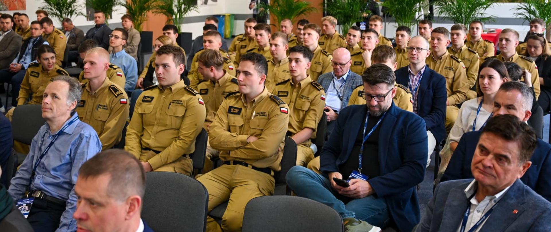 Strażak mówiąc do mikrofonu zabiera głos przed salą uczestników, którzy zajmują miejsca siedzące i słuchają wystąpień osoby ze sceny. W tle jest wyświetlana prezentacja.