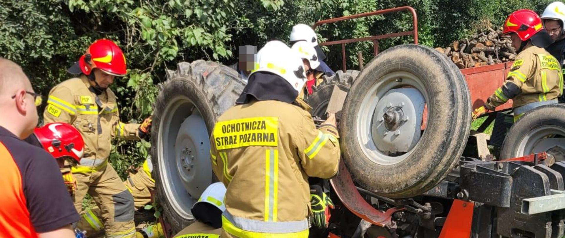 Zdjęcie przedstawia przewrócony ciągnik i strażaków, którzy unoszą ciągnik przy pomocy poduszek pneumatycznych.