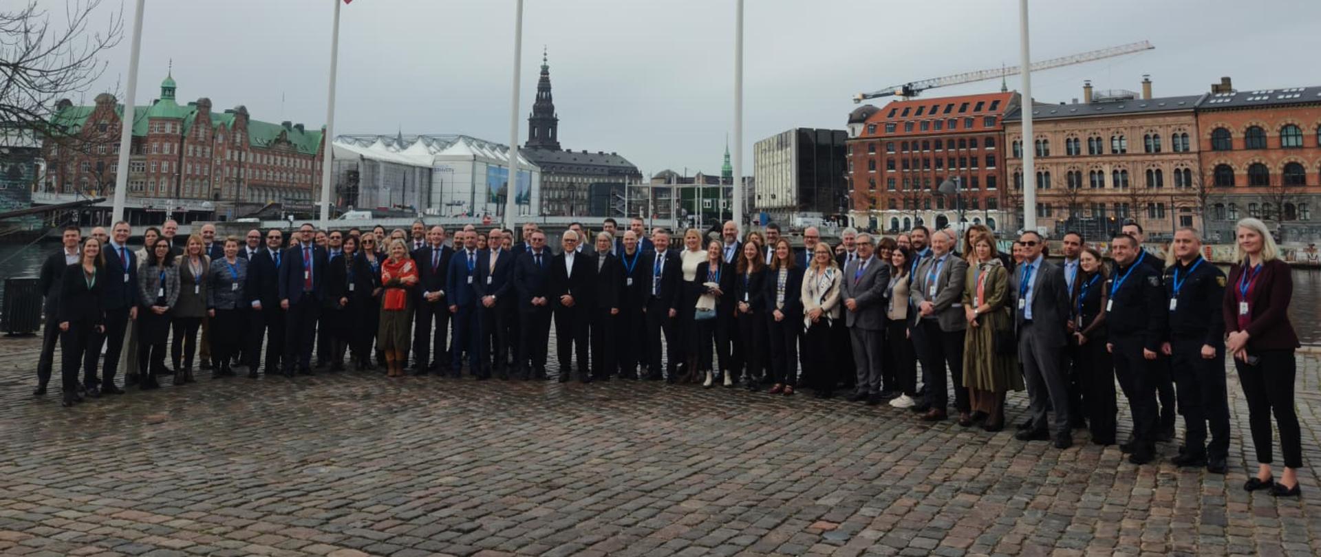 Grupa osób stoi na placu z brukowaną nawierzchnią, w tle widoczne są budynki o klasycznej architekturze i wieża z zegarem. Na pierwszym planie znajdują się cztery wysokie maszty z flagami Unii Europejskiej i Danii.
