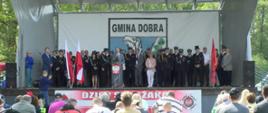 Na jasnej, zadaszonej scenie stoi kilkudziesięcioosobowa grupa osób, gospodarze uroczystości oraz zaproszeni goście. Część osób w mundurach. W centralnym miejscu przeźroczysta mównica z polskim godłem. W tle fragment logotypu z napisem Gmina Dobra. Na dole zdjęcia duży baner z napisem Dzień Strażaka i głowy siedzących na widowni dużej grupy osób.