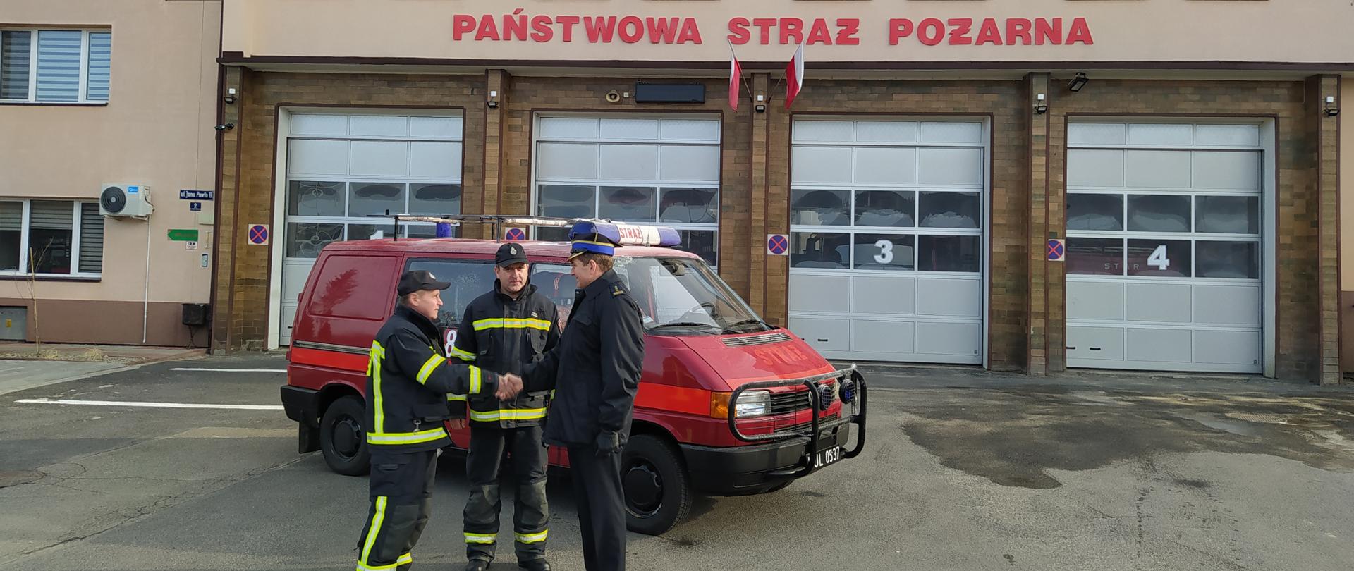 Zdjęcie przedstawia Komendanta Powiatowego Państwowej Straży Pożarnej w Lesku przed budynkiem Komendy Powiatowej Państwowej Straży Pożarnej w Lesku w mundurze wyjściowym przy samochodzie SLKw Volkswagen Transporter T4. Komendant Powiatowy Państwowej Straży Pożarnej w Lesku podczas uścisku dłoni z jednym z przedstawicieli Ochotniczej Straży Pożarnej w Średniej Wsi, przemawia. Dwóch druhów na zdjęciu ubranych jest w ubrania specjalne.