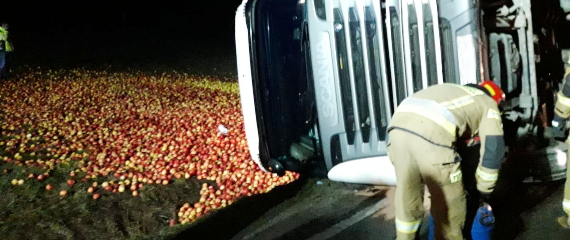 Samochód ciężarowy leżący na boku a na łące rozsypane jabłka. Obok samochodu strażak.
