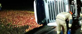 Samochód ciężarowy leżący na boku a na łące rozsypane jabłka. Obok samochodu strażak.