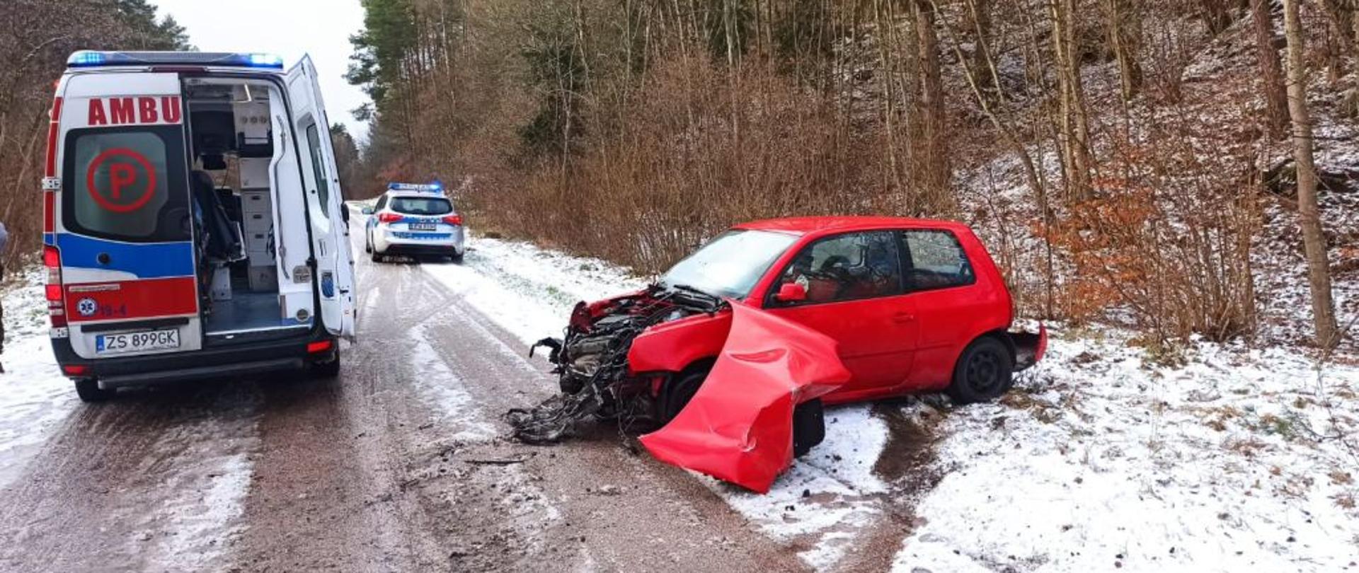 Widok na czerwone, małe auto osobowe. Auto ma rozbity przód. Przy aucie karetka pogotowia.