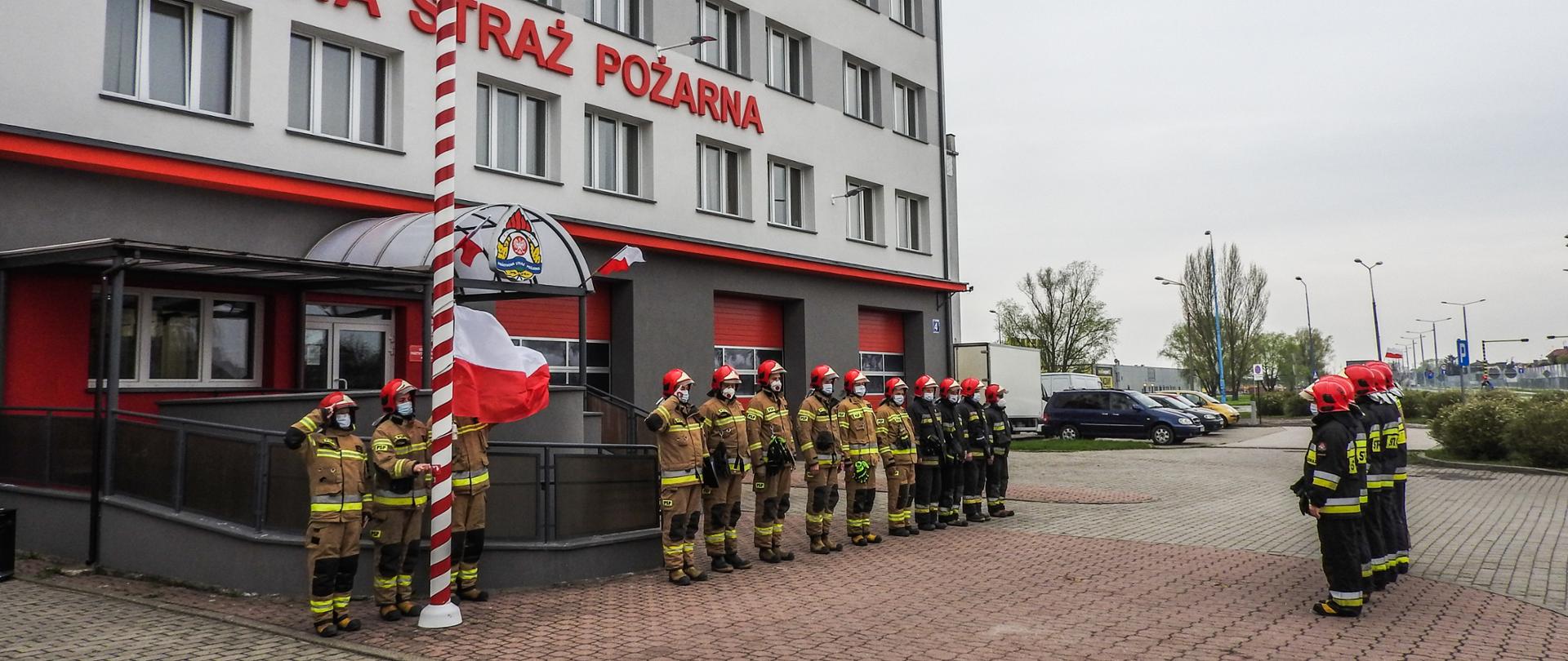 Zdjęcie przedstawia uroczystą zmianę służby w dniu 2 maja 2021 r. Podczas zmiany służby przed budynkiem komendy podniesiono flagę Polski i odegrano hymn państwowy. Strażacy występujący podczas zmiany ubrani byli w ubrania specjalne.