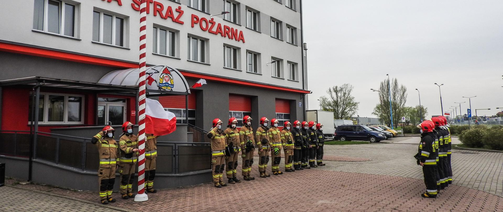 Zdjęcie przedstawia uroczystą zmianę służby w dniu 2 maja 2021 r. Podczas zmiany służby przed budynkiem komendy podniesiono flagę Polski i odegrano hymn państwowy. Strażacy występujący podczas zmiany ubrani byli w ubrania specjalne.