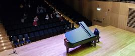 Widok z góry na estradę sali koncertowej, na środku której mężczyzna gra na fortepianie. W tle widać publiczność siedzącą na widowni.