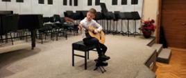 Chłopiec siedzi na krześle na środku estrady auli PSM i gra na gitarze. Za nim, pod ścianą stoją krzesła i pulpity oraz z lewej strony fortepian.