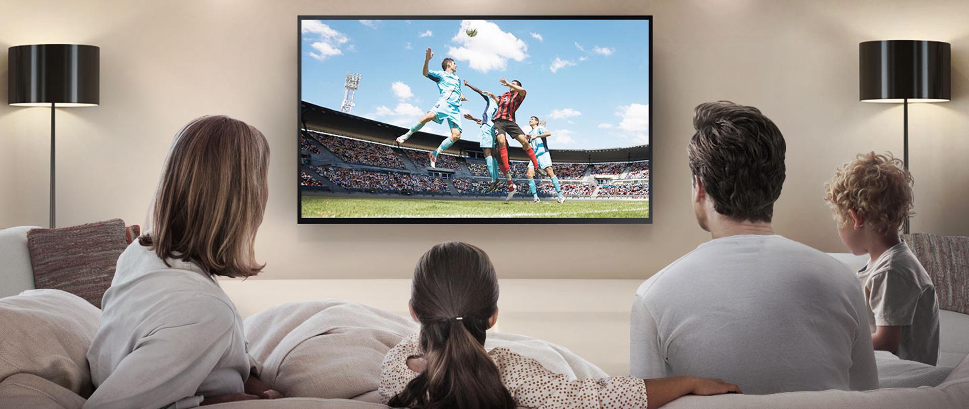 Zdjęcie ilustracyjne w poziomie. Na pierwszym planie biała kanapa, na której siedzi rodzina. Na dalszym planie telewizor, na którym wyświetlany jest mecz piłki nożnej.