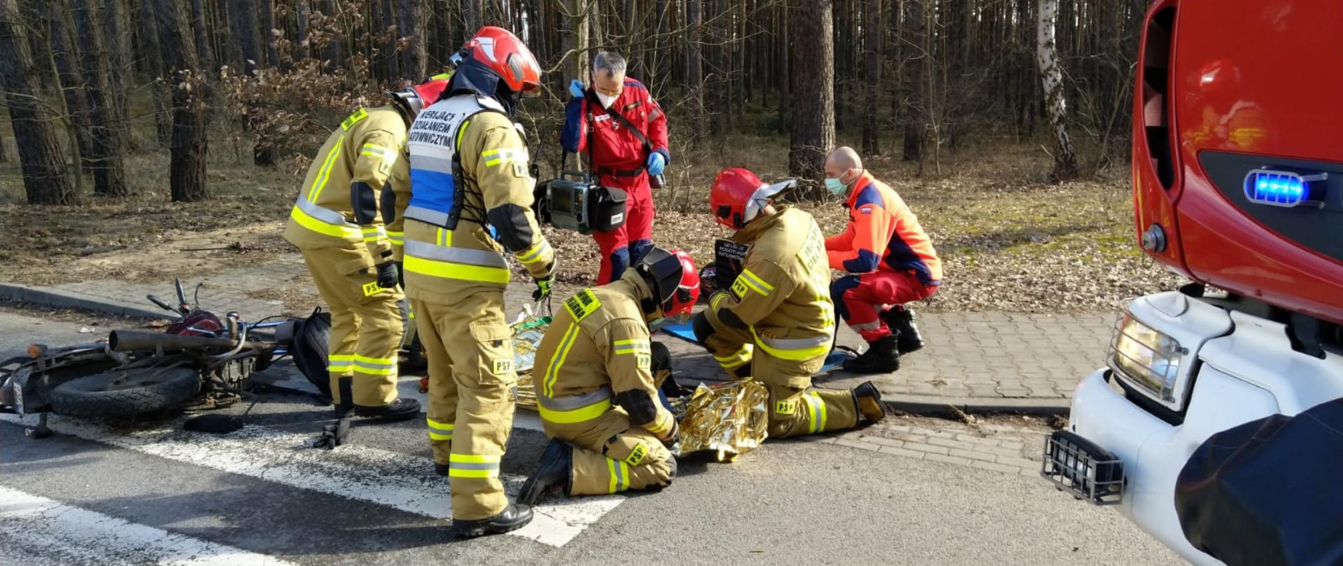 strażacyc wraz z ratownikami medycznymi udzielają pomoc poszkodwanemu w wypadku motocykliście