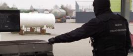 Funkcjonariusz KAS stoi przy dystrybutorze paliwa, w tle zbiornik na gaz LPG oraz cysterna