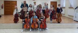 Fotografia przedstawia uczniów klasy wiolonczeli stojących z wiolonczelami oraz Panią Annę Skibińską i Urszulę Właźniak, które stoją razem z nimi. W tle brązowe drzwi, na górze wisi Godło Polski, w prawym rogu widać okna i białe firanki. 