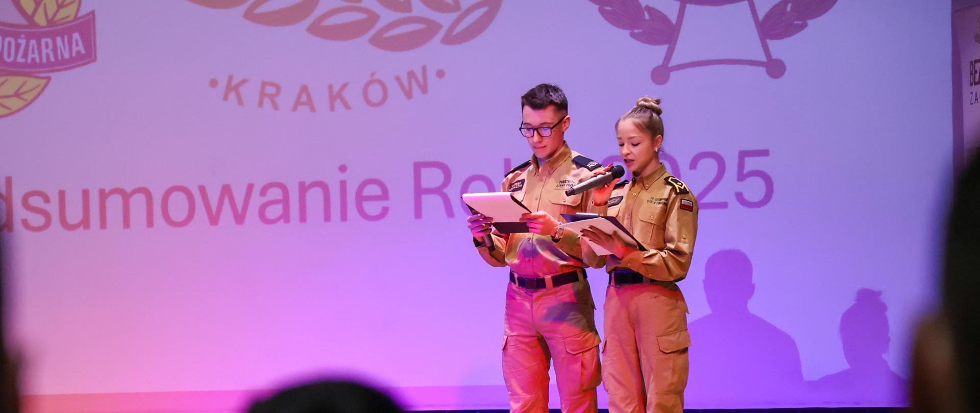 Dwoje młodych funkcjonariuszy (kobieta i mężczyzna) w piaskowych mundurach służbowych stoi na scenie przed dużym ekranem z logo PSP. Czytają tekst z dokumentów, prowadząc oficjalną część spotkania.