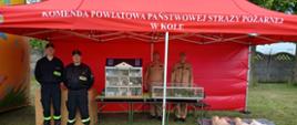 Czterech strażaków stoi pod czerwonym namiotem z napisem "Komenda Powiatowa Państwowej Straży Pożarnej w Kole". Przed nimi znajdują się modele budynków i manekiny do nauki resuscytacji. Dwóch strażaków ma czarne mundury z napisem "STRAŻ", a dwóch beżowe mundury PSP.