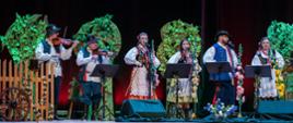 Na scenie osoby ubrane w stroje ludowe grają na skrzypcach i śpiewają. Obok sceneria wiejska: płotek, rośliny, kwiaty.