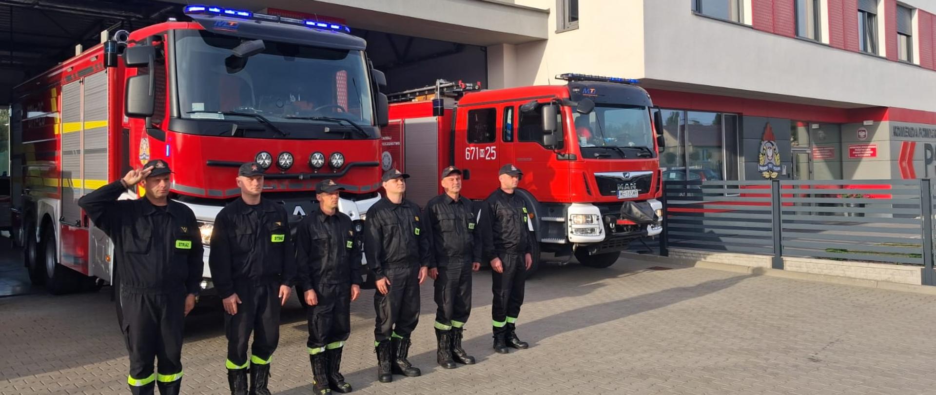 Strażacy stoją na baczność, dowódca z lewej salutuje. Wszyscy stoją na tle samochodów pożarniczych oddając tym samym hołd zmarłemu strażakowi z Kalwy.