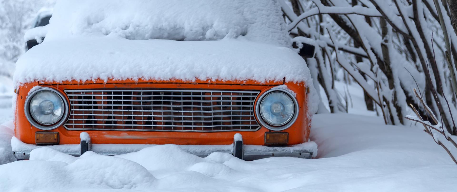 Pomarańczowy samochód stojący i zasypany śniegiem - widoczny jest tylko jego przód.