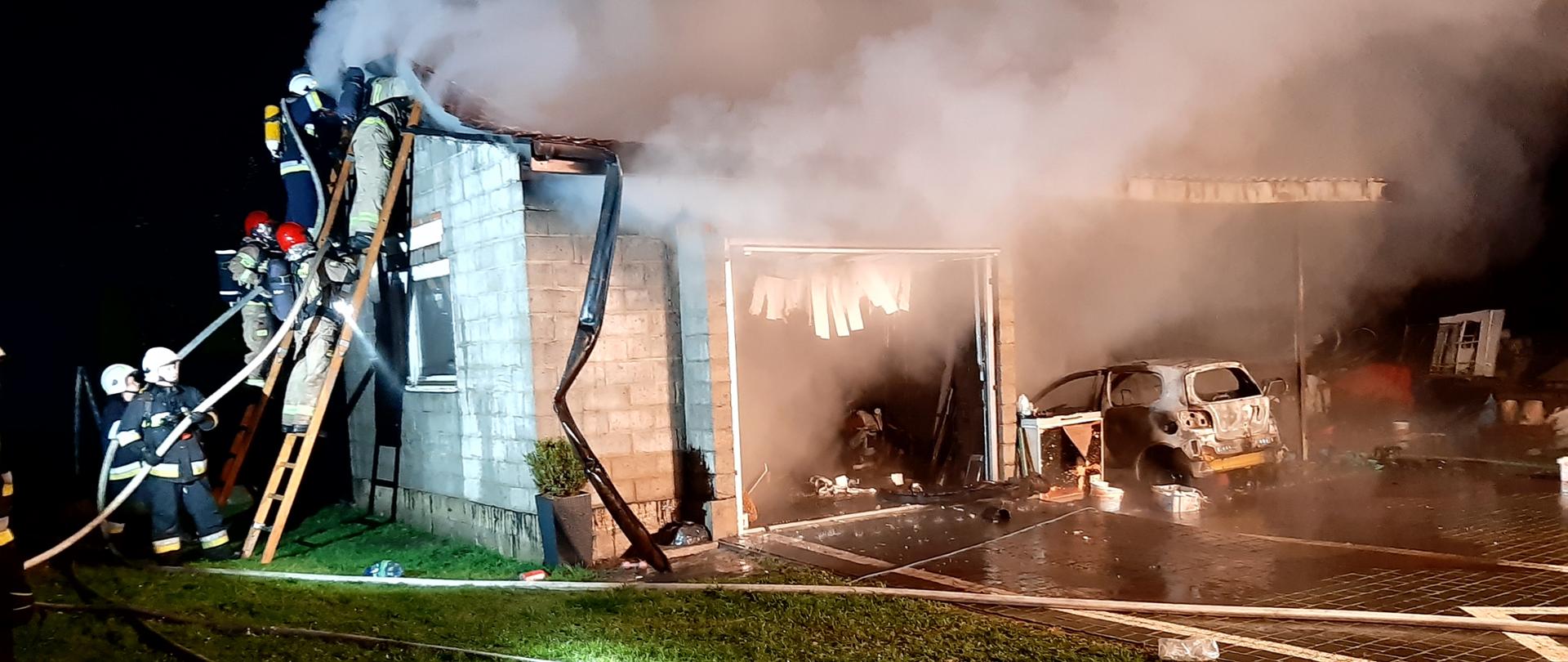 Zdjęcie przedstawia garaż wolnostojący w trakcie pożaru. Dwóch ratowników gasi pożar z drabiny dostawnej, którą zabezpiecza dodatkowych 4. W jednej z bram widać spalony samochód osobowy.
