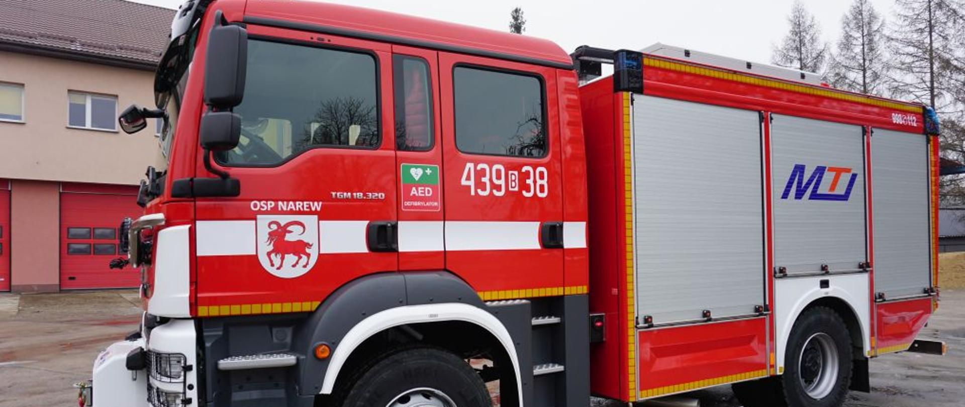 Strażacki wóz bojowy koloru czerwonego z napisem OSP Narew. Z boku samochodu trzy zamknięte żaluzje.