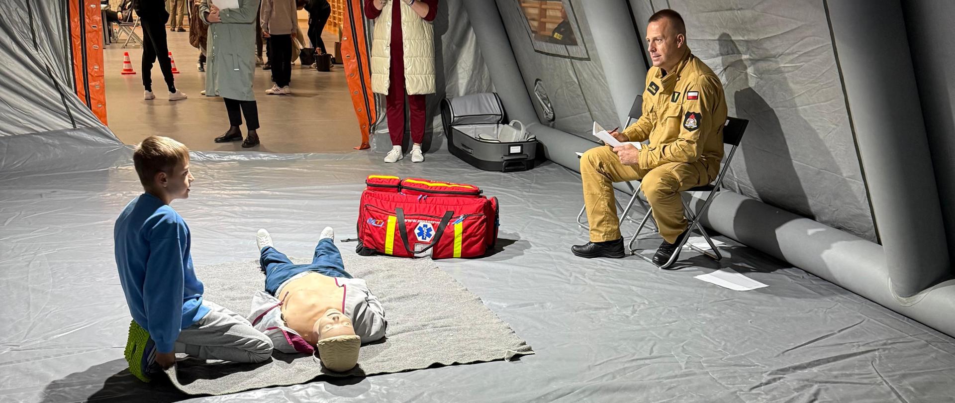Wewnątrz dużego, dmuchanego namiotu ratowniczego odbywa się praktyczne szkolenie z udzielania pierwszej pomocy, podczas którego młody chłopiec ćwiczy resuscytację na manekinie, pod okiem instruktora w mundurze siedzącego po prawej stronie. Scenę, w której kluczowe są manekin i czerwona torba medyczna, obserwuje w wejściu do namiotu niewielka grupa osób.