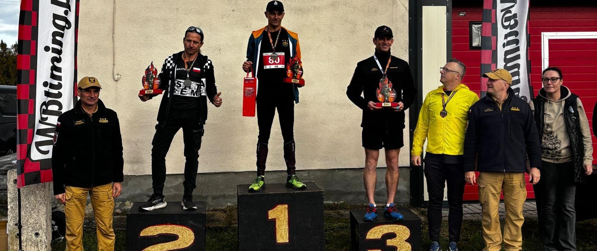 Na tle ściany budynku stoi podium ze sportowcami. Po bokach strażacy i organizatorzy zawodów sportowych.