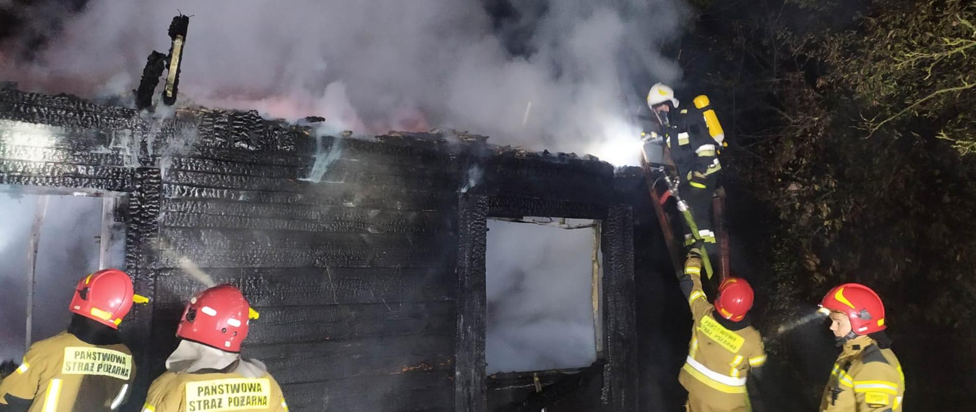 widoczna drewniana ściana budynku, cała zwęglona, przy drzwiach stoi dwóch strażaków, podają wodę do środka, na prawo od nich jeden stoi na drabinie sięga po sprzęt podawany mu przez drugiego, asekurującego drabinę, wewnątrz domu duże zadymienie