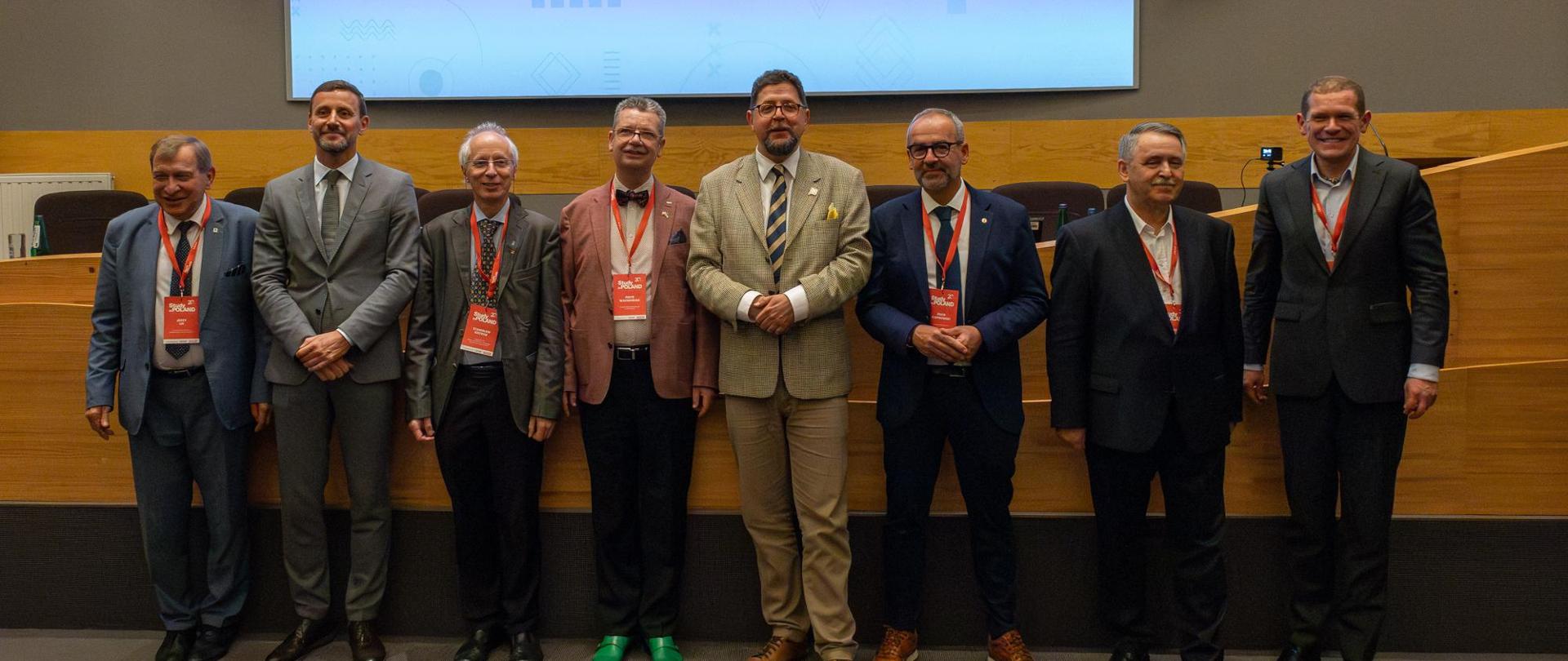 Zdjęcie zbiorowe, pod wyłożona drewnianymi panelami ścianą stoi dziewięciu mężczyzn w garniturach, pośrodku grupy wiceminister Szeptycki.