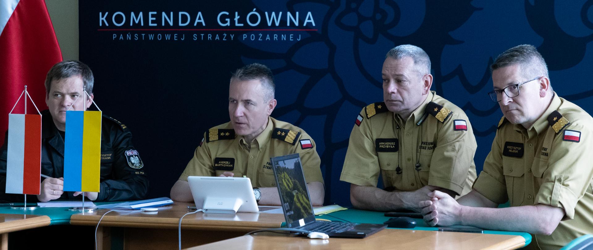 Na zdjęciu widać czterech strażaków podczas wideokonferencji. Po lewej stronie na stoliku małe flagi Polski i Ukrainy. W tle tło koloru ciemnoniebieskiego z napisem Komenda Główna Państwowej Straży Pożarnej.