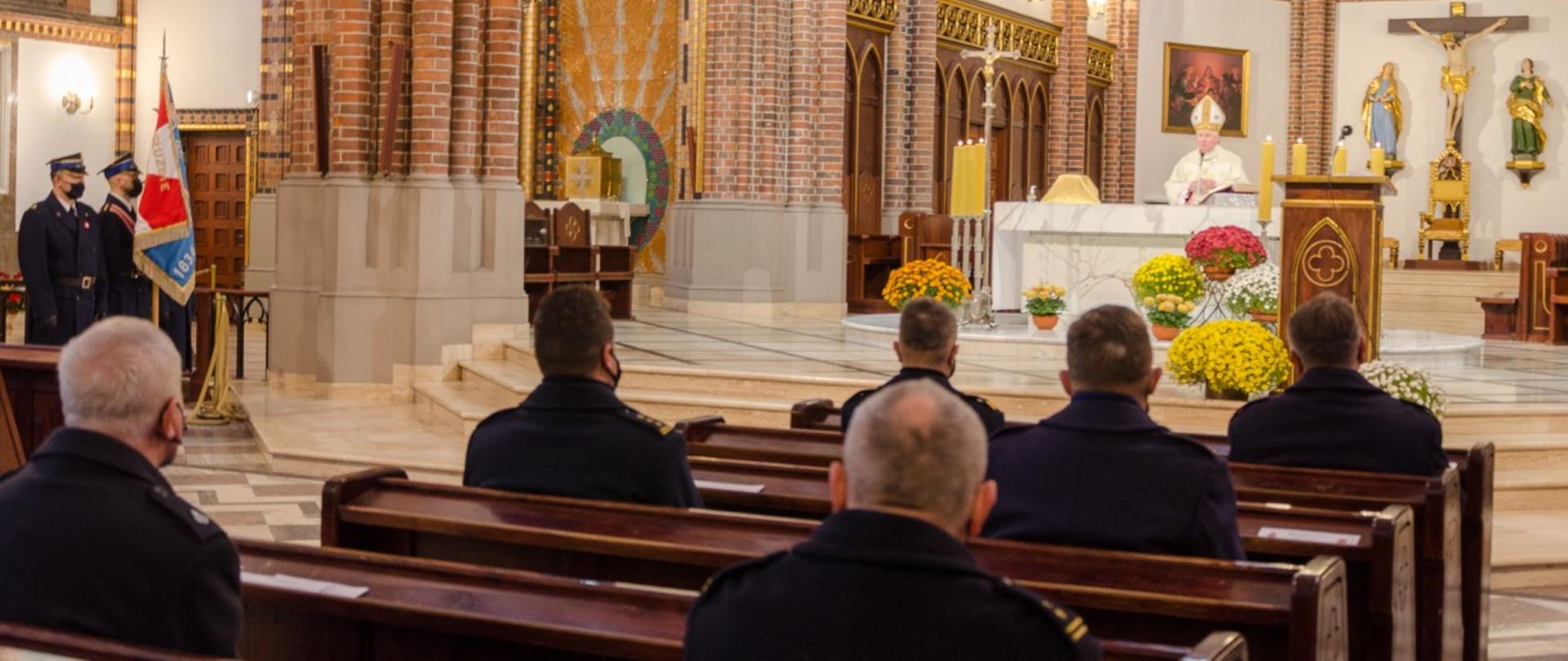 Zdjęcie przedstawia ołtarz w katedrze św. Floriana w Warszawie. Na pierwszym planie strażacy siedzą w ławkach. W głębi w centralnym miejscu ksiądz biskup odprawia nabożeństwo. Po lewej stronie zdjęcia stoi poczet sztandarowy Państwowej Straży Pożarnej.