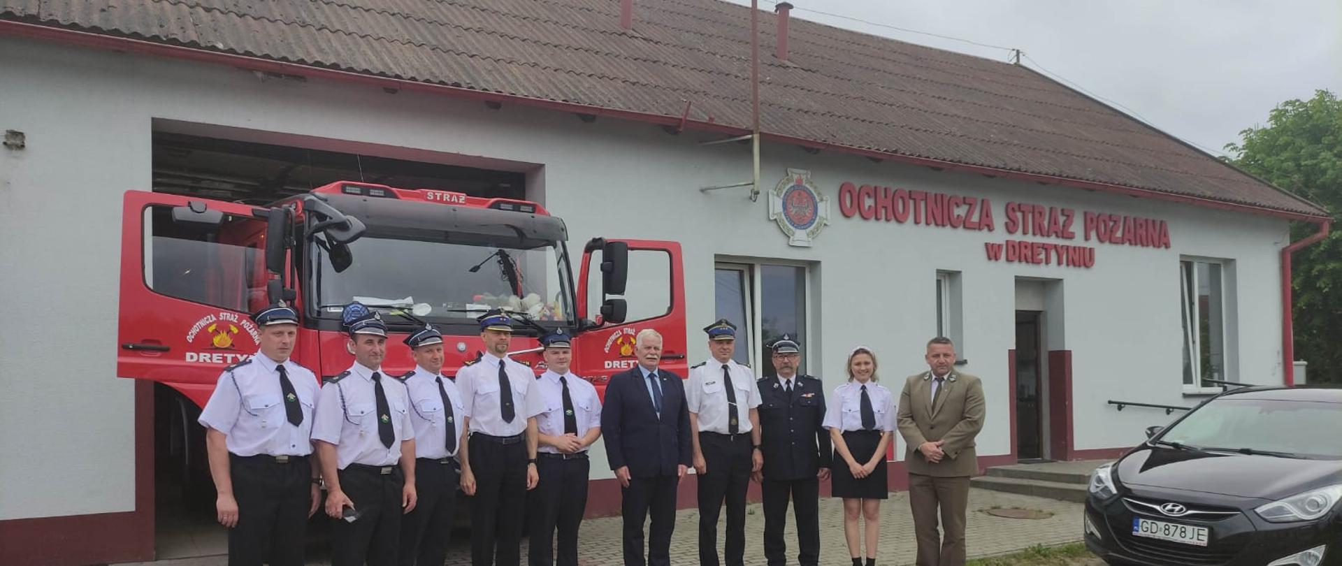 Na pierwszym planie zdjęcia widać osoby ubrane w mundury wyjściowe ustawione w szeregu na tle budynku strażnicy ochotniczej straży pożarnej w Dretyniu.