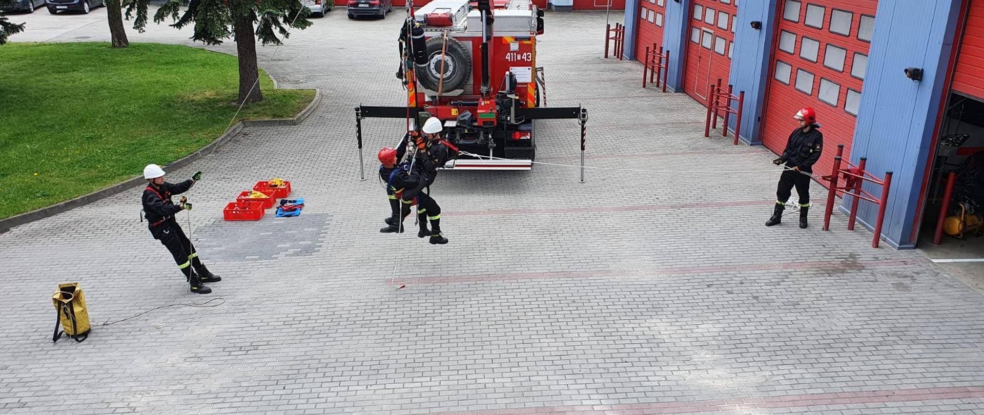 Zdjęcie przedstawia na pierwszym planie dwóch strażaków którzy zabezpieczeni w sprzęt wysokościowy znajdują się na pewnej wysokości ponad ziemią. Strażacy przypięci są hydraulicznego dźwigu samochodowego będącego na wyposażeniu specjalistycznego samochodu ratownictwa technicznego marki Scania. Po lewej stronie strażak asekuruje kolegów, natomiast po prawej stronie kolejny zabezpiecza ich.