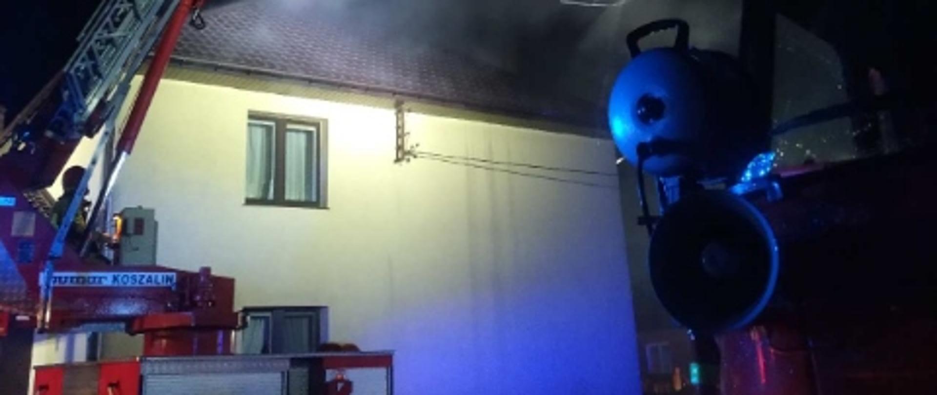 W środku zdjęcia budynek piętrowy, jednorodzinny w kolorze żółtym. Dach blaszany, brązowy. Nad dachem unosi się szary dym. Budynek oświetlony z najaśnicy. Po lewej podnośnik z JRG 2 Kalisz. Pojazd w kolorze czerwonym. Zdjęcie wykonane w porze nocnej. W prawym, dolnym rogu fragment samochodu gaśniczego star.