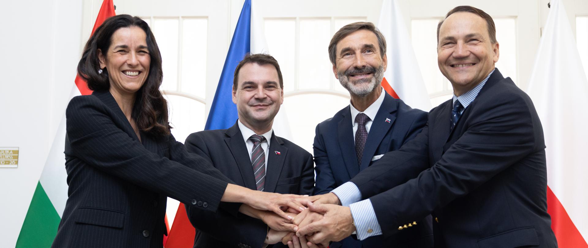 Ministrowie Węgier, Czech, Słowacji i Polski stoją przed flagami Polski, Węgier, Słowacji i Czech. Wszyscy wspólnie układają dłonie na środku w geście solidarności i współpracy; wszyscy uśmiechają się do aparatu.