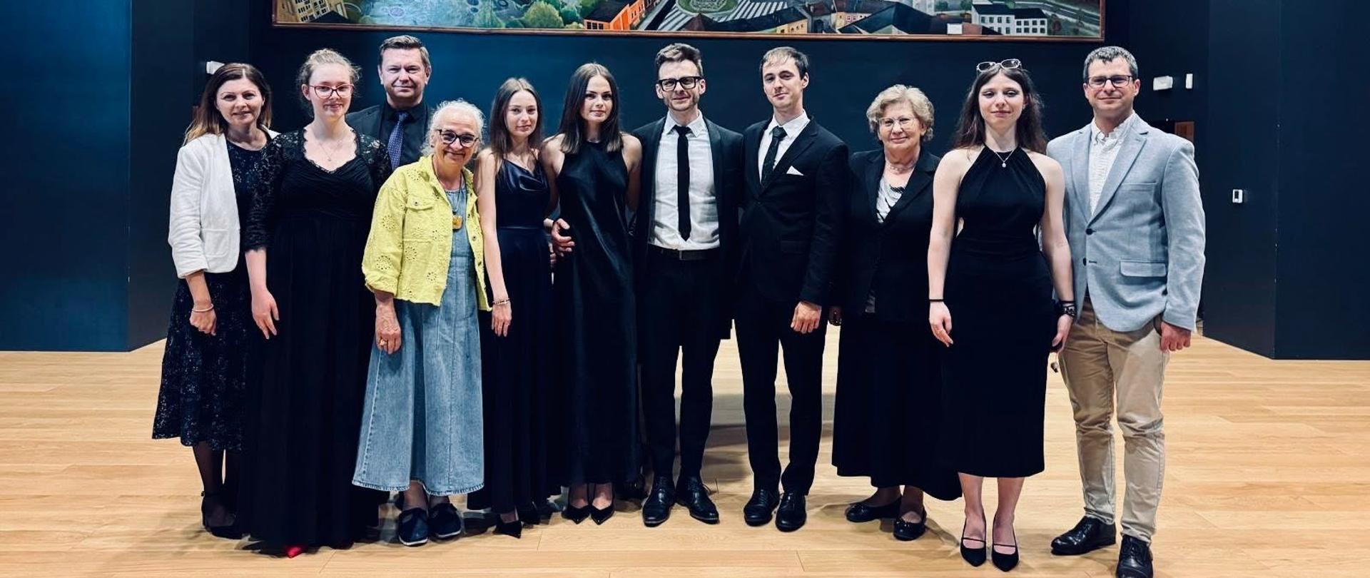 Dziesięć osób stoi w rzędzie na tle dużego obrazu przedstawiającego panoramę miasta z charakterystycznymi czerwonymi dachami i katedrą. Grupa składa się z muzyków i organizatorów ubranych elegancko - większość w czarne stroje, jedna osoba w żółtej marynarce, jedna w jasnym garniturze. Wnętrze ma nowoczesny charakter z ciemnoniebieskimi ścianami, profesjonalnym oświetleniem i drewnianą podłogą. Wszyscy uśmiechają się.