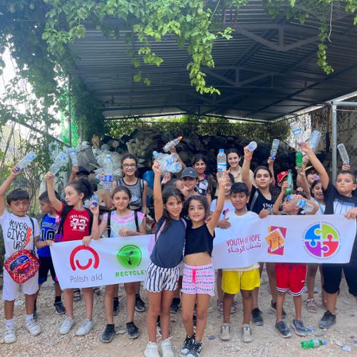 Na tle wiaty z odpadami plastikowymi stoi grupa uczniów trzymających w rękach banery i plastikowe butelki