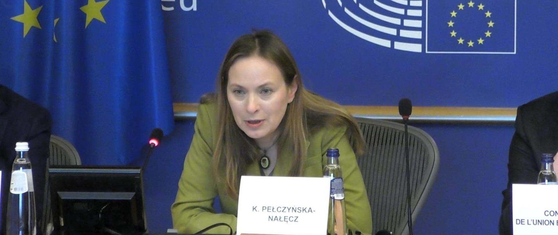 Minister Katarzyna Pełczyńska-Nałęcz siedzi za stołem i przemawia, za jej plecami granatowy baner przedstawiający Parlament Europejski i flaga UE