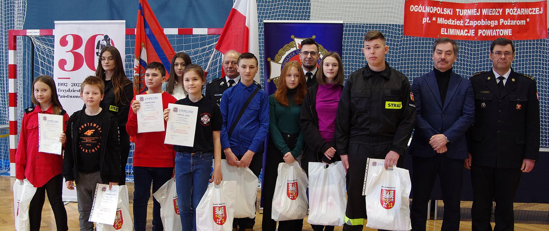 Widok z przodu. Zdjęcie zbiorowe finalistów OTWP z organizatorami i gośćmi. W tle flaga Polski i flaga Związku OSP RP oraz wiszący baner „Ogólnopolski Turniej Wiedzy Pożarniczej pt. Młodzież zapobiega pożarom – eliminacje powiatowe”.