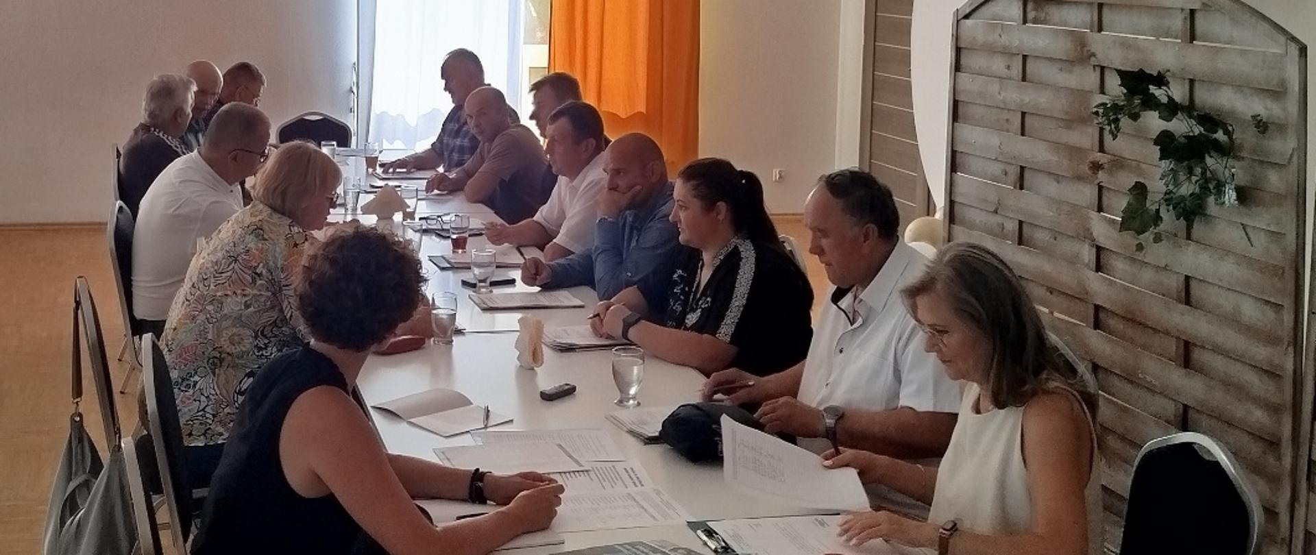 Grupa osób siedzi przy długim stole, uczestnicząc w spotkaniu i przeglądając dokumenty.