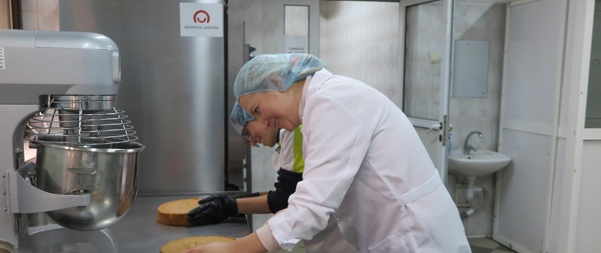 Dwie kobiety przygotowują ciasta w przemysłowej kuchni, w tle widać naklejkę z logo Polska pomoc