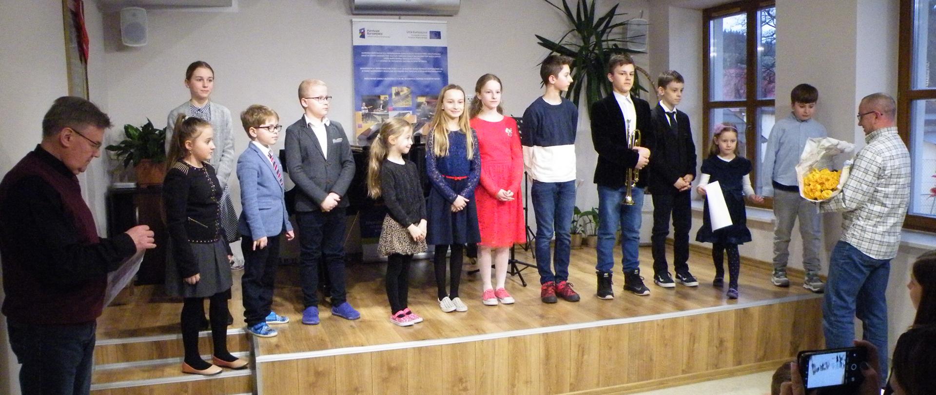 Na sali koncertowej na scenie uczniowie biorący udział w koncercie z okazji dnia kobiet. W tle fortepian, pulpity oraz baner informujący o funduszach europejskich. Po prawej stronie zielona wysoka roślina. Na pierwszym planie po lewej stronie stojący kierownik filii a po prawej stronie trzymający w ręku kwiaty rodzic