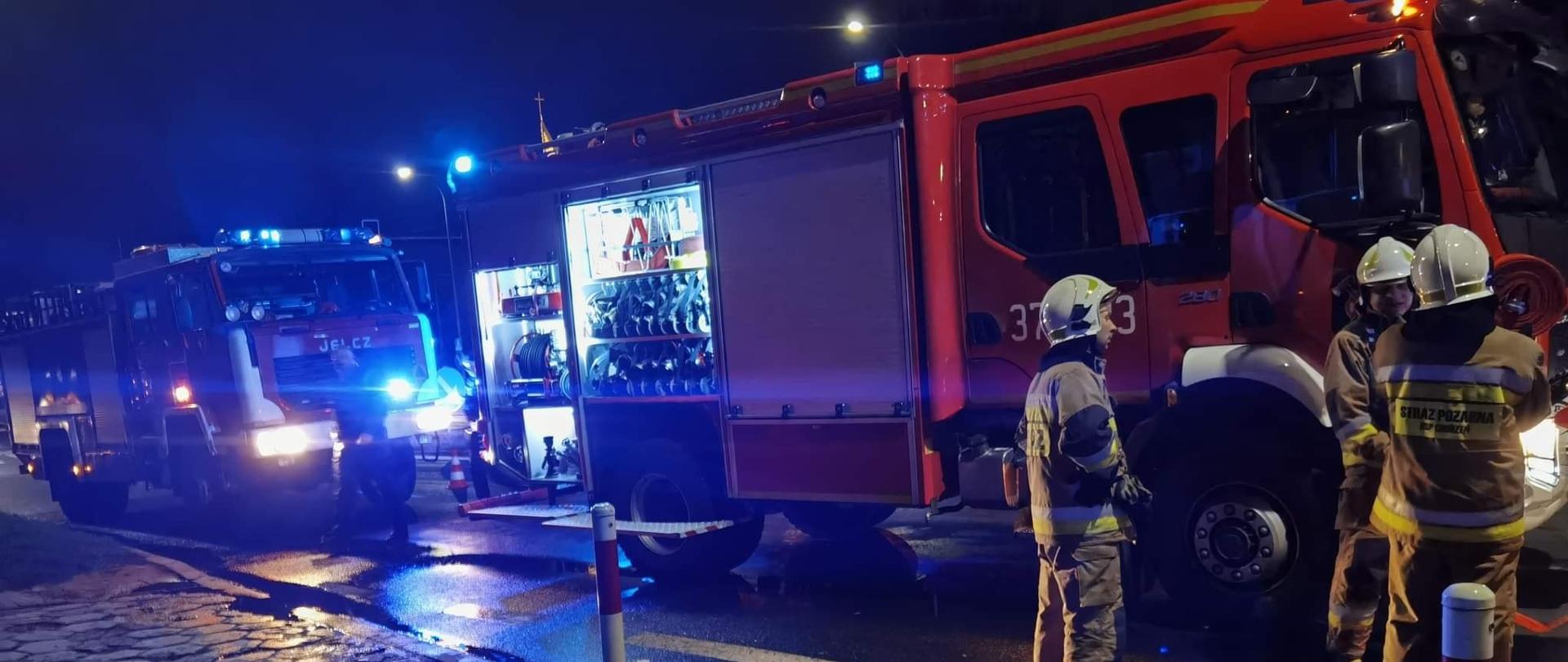 Zdjęcie przedstawia dwa samochody strażackie stojące na ulicy. Przed samochodami stoją strażacy. Pora nocna. Samochody mają włączone sygnały świetlne. 