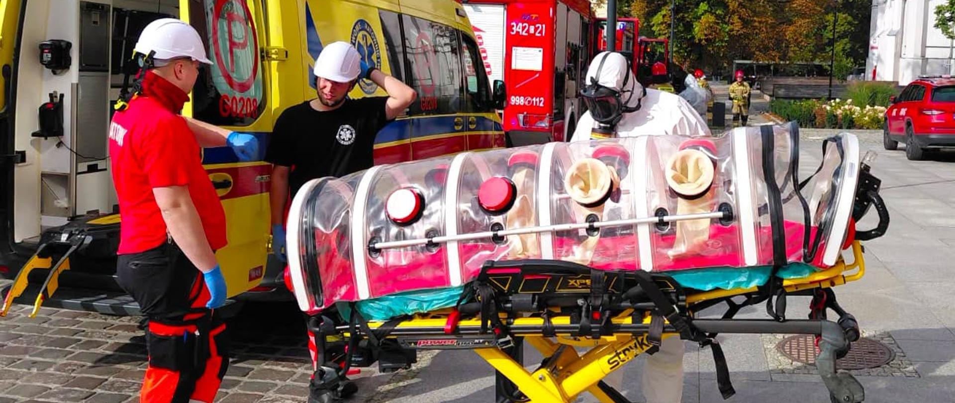Zdjęcie przestawia osobie znajdująca się na noszach, zamkniętą ochronnej foli. Obok ratownik medyny oraz osoba ubrana w ubranie chony chemicznej. 