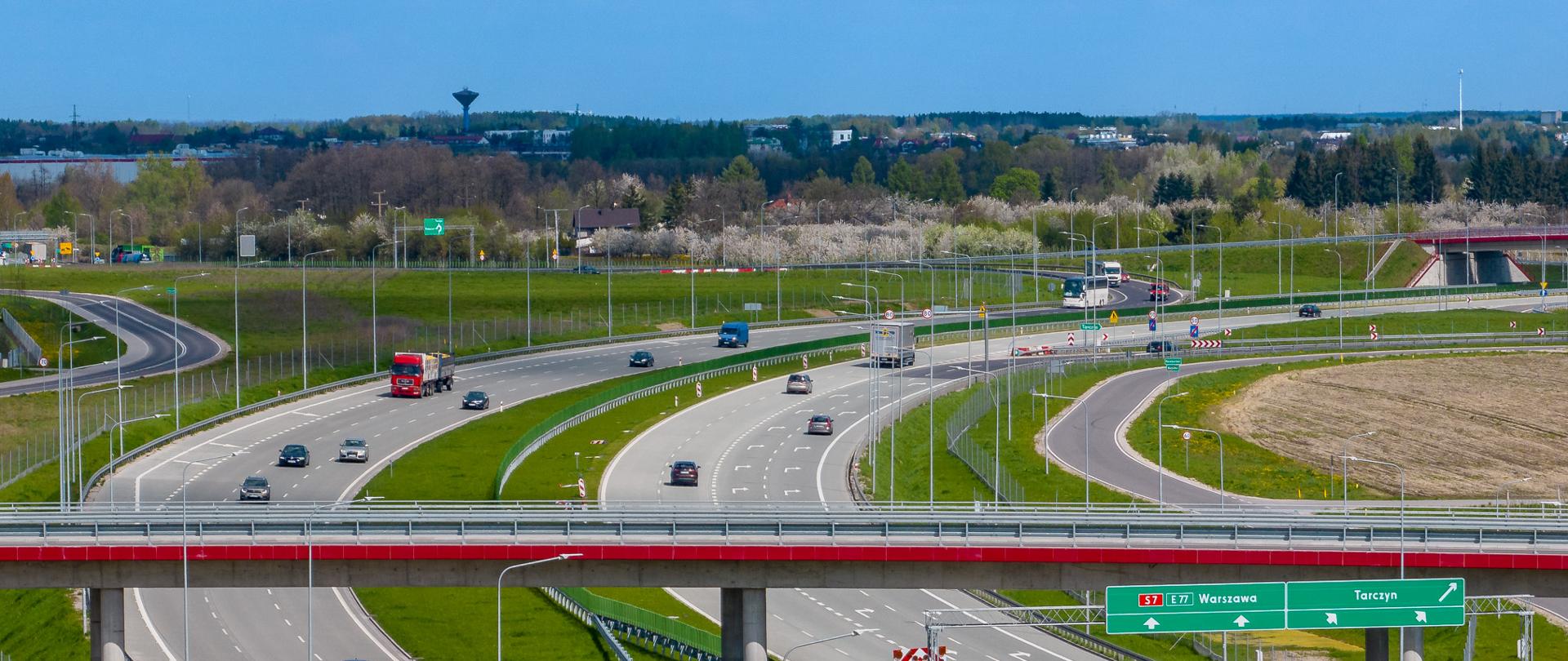 Na zdjęciu widać ruchliwą drogę ekspresową z wieloma pasami ruchu w obu kierunkach. Pojazdy, w tym ciężarówki i samochody jadą główną trasą, włączają się do ruchu lub zjeżdżając z trasy. W centrum widać wiadukt i znaki drogowe wskazujące kierunki. Niebo jest bezchmurne. Wokół drogi rozciągają się zielone łąki i zaorane pola uprawne.
