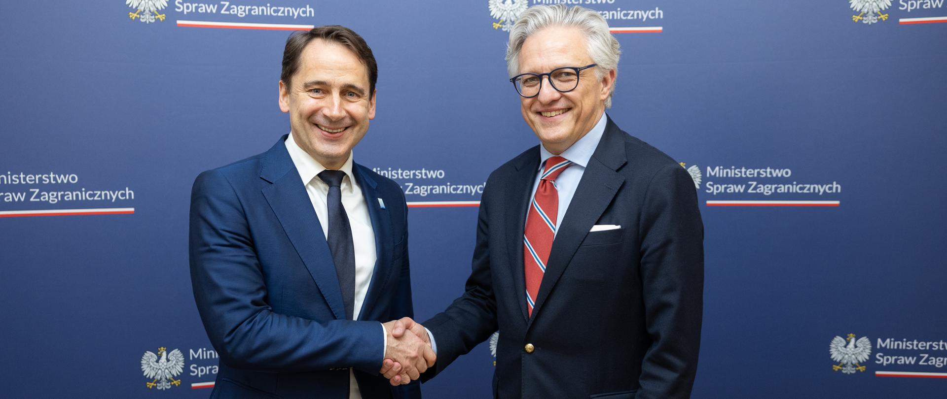 Podsekretarz Stanu w MSZ Artur Harazim i Wiceminister Spraw Zagranicznych Litwy Sigitas Mitkus uśmiechają się podczas uścisku dłoni na tle granatowej ścianki z logo Ministerstwa Spraw Zagranicznych 