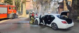 Na zdjęciu widać palący się biały samochód osobowy oraz dwóch strażaków podczas akcji gaśniczej. W tle widoczny jest samochód ratowniczo gaśniczy.